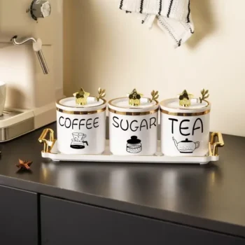Tre enë dekorative plastike me mbishkrime Coffee, Sugar dhe Tea, të vendosura në tabaka të bardhë me detaje të arta.
